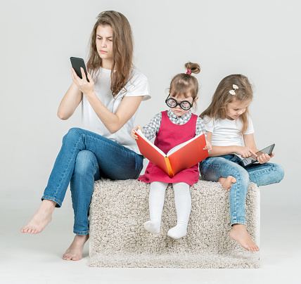 Three cute sisters are sitting together. The teenager and middle preschooler dressed in a t-shirts and in jeans, the younger girl in casual dress. The older sister and the middle sister are using the digital gadgets and browsing the internet. The younger sister is reading the book. Studio shooting