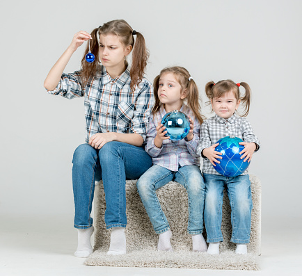 Three cute sisters (teenager and two preschoolers) dressed in casual clothing are sitting together. The girls are holding the various blue balls in hands (christmas balls). The sisters are showing the different emotions, the older sister is disappointed and dissatisfied, the middle sister is surprised and the younger sister is very happy. Studio shooting