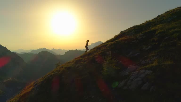 AERIAL SILHOUETTE: Contour of young woman walking up mountain at golden lit dusk