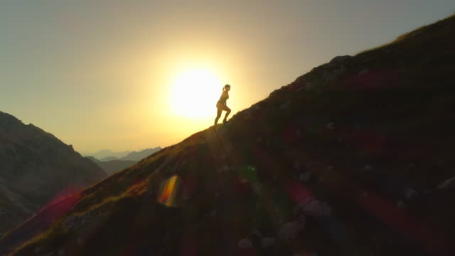 AERIAL SILHOUETTE: Woman trekker trying to reach mountain peak before sundown.