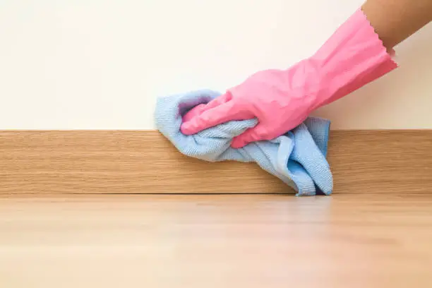 Photo of Employee hand in rubber protective glove with micro fiber cloth wiping a baseboard on the floor from dust at the wall. Spring general or regular clean up. Commercial cleaning company concept.