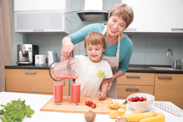 幸せな祖母と小さな孫がキッチンでミキサーで新鮮なフルーツとベリーのスムージーを作るします。小さな男の子は、朝食にフルーツ カクテルやスムージーの準備します。健康的な食事 - two pears ストックフォトと画像