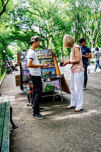Visitor paying for fridge magnets from a vendor on Literary Walk, also known as The Mall, in Central Park, Manhattan, New York City, on an overcast summer's day.