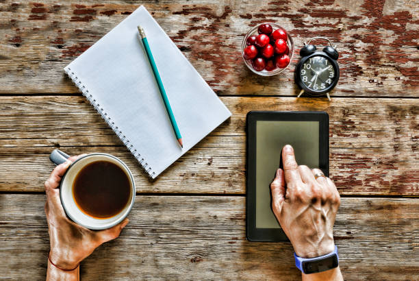 Freelancer works with a tablet computer at home at the table, next to it there is an open notepad and a pencil, a clock. In his hand a cup of coffee. The concept: work outside the office, freelancing, remote work.