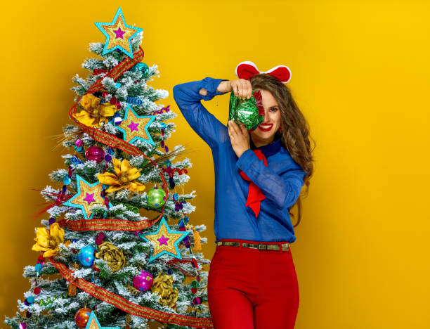 Festive season. happy modern woman near Christmas tree on yellow background with wrapped as a gift photo camera