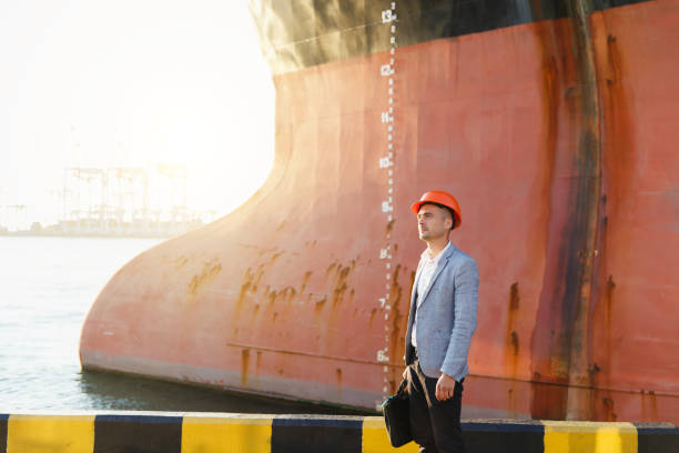 istock Handsome young unshaven successful business man in gray suit and protective construction orange helmet holding case, standing in sea port against a cargo rusty ship with water line background 880508086