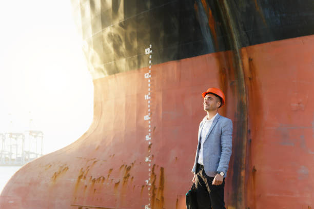 istock Handsome young unshaven successful business man in gray suit and protective construction orange helmet holding case, standing in sea port against a cargo rusty ship with water line background 880508138