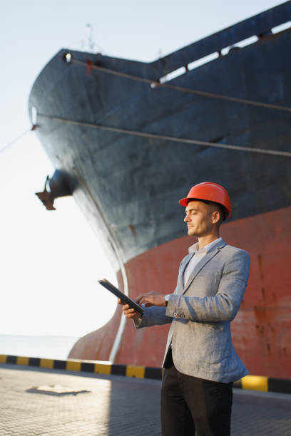 Handsome unshaven successful business man in gray suit, protective construction orange helmet holding tablet, standing in sea port against cargo rusty ship background. Male with gadget in sunny day Handsome unshaven successful business man in gray suit, protective construction orange helmet holding tablet, standing in sea port against cargo rusty ship background. Male with gadget in sunny day. business architecture blue people stock pictures, royalty-free photos & images