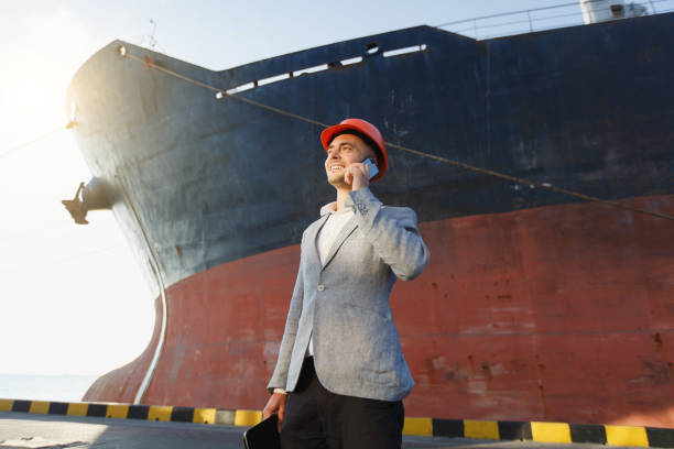 istock Handsome young unshaven successful business man in gray suit and protective construction orange helmet holding tablet, talking on mobile phone, standing in sea port against cargo rusty ship background 880508696