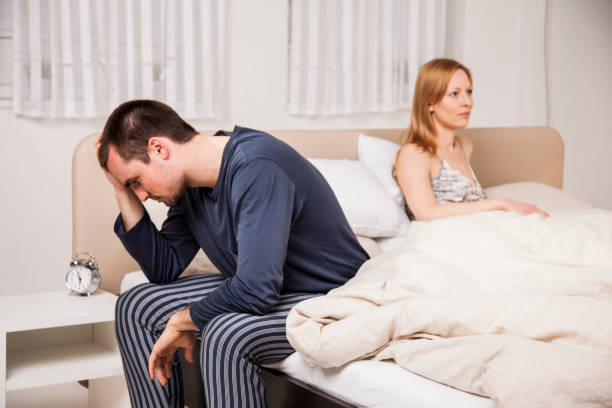 Couple with problems Young couple in bed. Man with hand on his head and sitting on the side of bed. Woman lying on bed facing other direction. Having problems. sti stock pictures, royalty-free photos & images