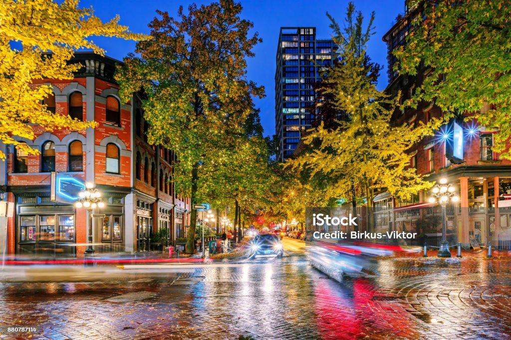 Night on the street in the old part of the big city Night on the street in the old part of the big city with the lights of storefronts and shops, the blinding lights of car headlights and a paved roadway shimmering in neon lights after the last rain Vancouver - Canada Stock Photo