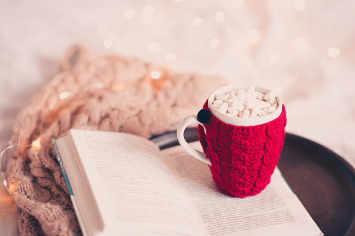 Knitted cup of coffee on open book with knitted scarf in bed over Christmas lights closeup. Good morning.