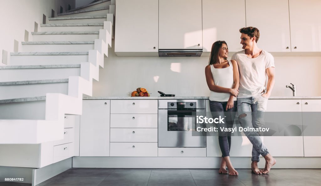 Couple at home. Full-length image of romantic couple at home. Attractive young woman and handsome man are enjoying spending time together while standing on light modern kitchen. Couple - Relationship Stock Photo