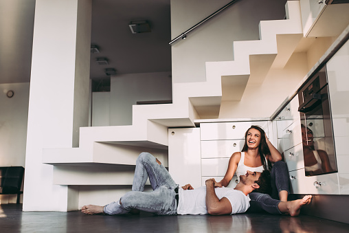 Romantic couple at home. Attractive young woman and handsome man are enjoying spending time together. Sitting on the floor in light modern kitchen.