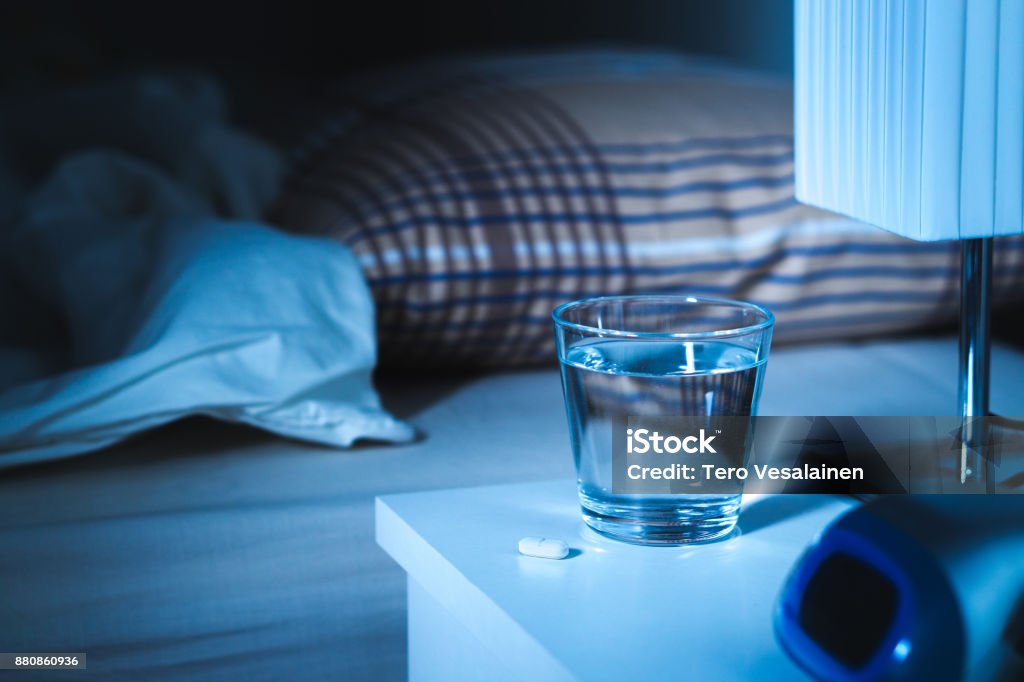 Sleeping pill on nightstand next to a glass of water. White tablet on table and bed in bedroom. Sleeping pill on nightstand next to a glass of water. White tablet on table and bed in bedroom. Insomnia medicine concept. Sleeping Pill Stock Photo