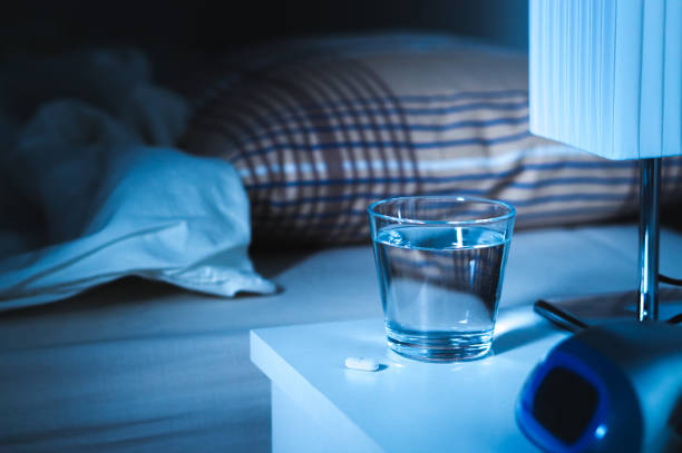 Sleeping pill on nightstand next to a glass of water. White tablet on table and bed in bedroom. Sleeping pill on nightstand next to a glass of water. White tablet on table and bed in bedroom. Insomnia medicine concept. sleeping pill stock pictures, royalty-free photos & images