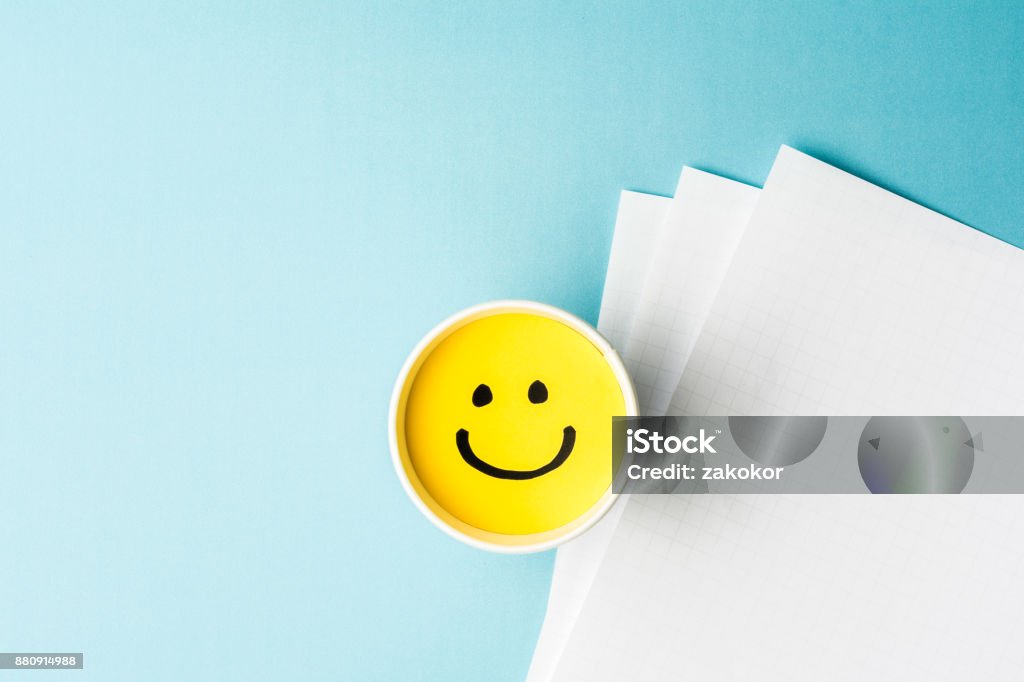 Yellow smiling face, happy mood, on paper cup and papers over blue background. Wellbeing Stock Photo