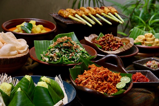 Nasi Campur Bali, the Balinese popular rice dish. Steamed turmeric rice served with side dishes such as Jukut Urap (vegetable salad with grated coconut dressing), Ayam Pelalah (shredded chicken in red chili paste), Telur Dadar (egg omelet), Kacang Teri (fried peanuts and anchovies), Sate Lilit (minced seafood satay), Kerupuk Udang (prawn crackers), Jukut Gedang Mekuah (green papaya curry) and condiments of Sambal Tomat (red chili and tomato paste) and Sambal Matah (chili and lemongrass salsa). The dishes are arranged on a table lined with traditional Balinese woven textile called Seseh cloth.