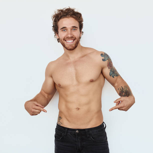 Man pointing at his abs Handsome young man pointing at his abs and smiling for camera. chest tattoo men stock pictures, royalty-free photos & images