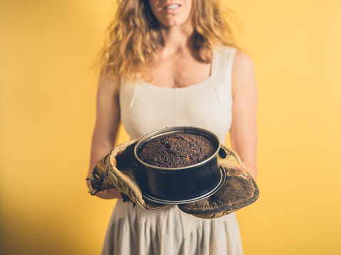 A young woman is holding a burnt cake with burnt oven gloves
