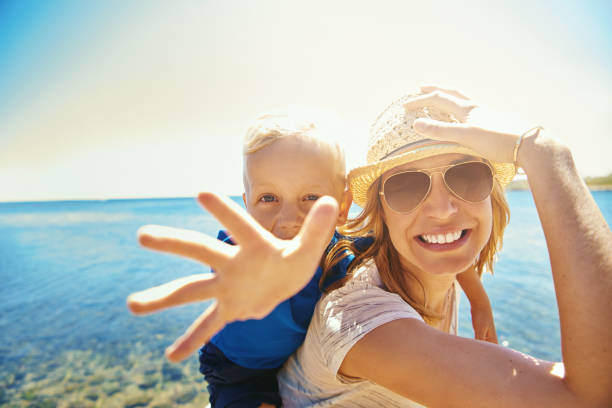 Young smiling mother with little boy on back that outstretching hand at camera on background of seaside.