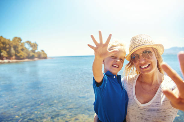 Cute little boy waving at the camera on a beach as she is held in his smiling mothers arms against a backdrop of the ocean