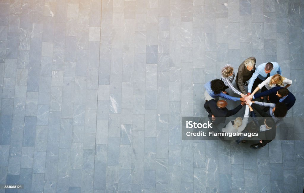 United colleagues standing in a huddle with their hands together High angle view of a team of united businesspeople standing with their hands together in a huddle in the lobby of a modern office building People Stock Photo