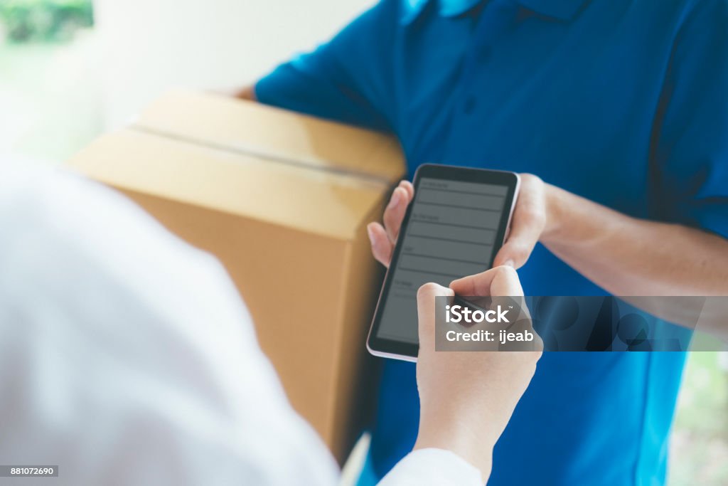 Woman signing on digital device to delivery parcel. Young woman appending signature in digital mobile phone after receiving parcel from courier at home. Delivering Stock Photo