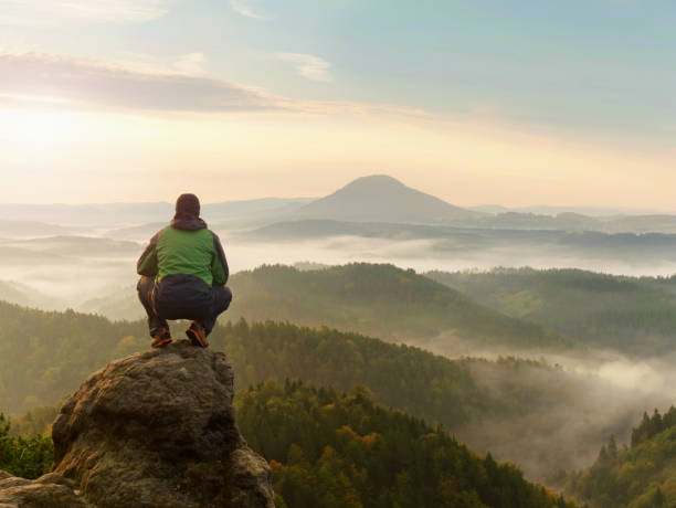 istock Man stay on rocky peak within daybreak and watch over misty landscape. 881147154
