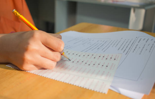 Students hands taking exams, writing examination room with holding pencil on optical form of standardized test with answers and english paper sheet on row desk chair doing final exam in classroom. stock photo