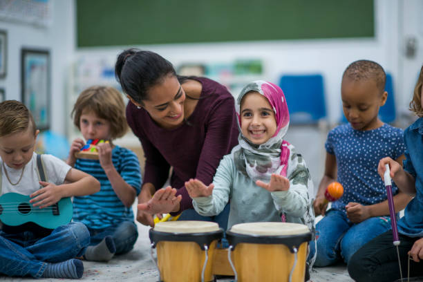 Girl Playing Drums A multi-ethnic group of young school children are indoors in their classroom. Their teacher is watching a girl playing drums. Her classmates are playing a flute, a recorder, a guitar, and maracas. rattle drum stock pictures, royalty-free photos & images