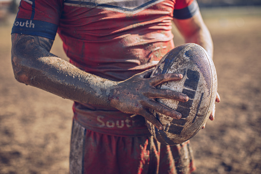One man, rugby player covered in mud, holding rugby ball in his nads, part of.\n