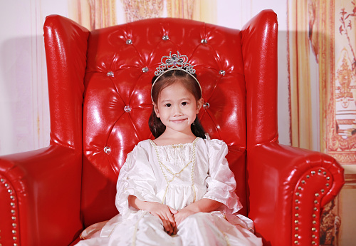 Pretty little girl in beautiful luxurious dress sitting on the red sofa.