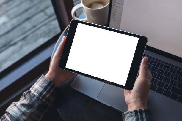 istock Mockup image of hands holding black tablet pc with white blank screen and laptop , coffee cup on wooden table background in cafe 881359542