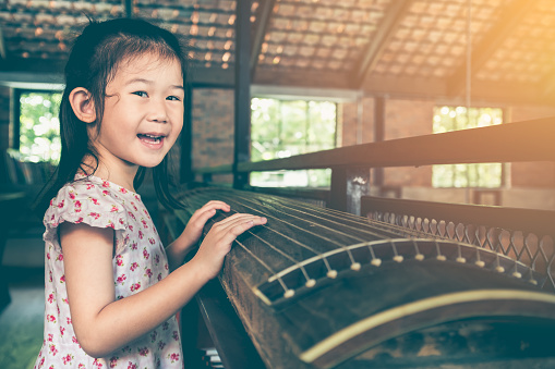 Attractive happy asian girl in beautiful dress smiling and looking at camera in her home. Child playing stringed instruments and having fun. Vintage film filter effect.