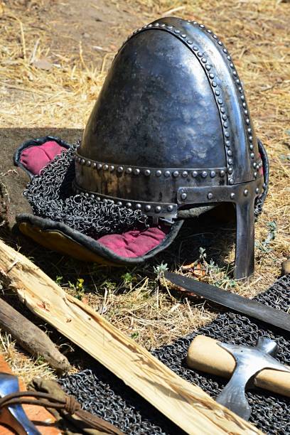 istock Medieval norman type conical helmet with nose protection piece and sides protected with chainmail placed on fabric filler next to battle axe, sword and chainmail armor 881381374