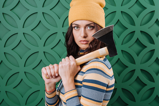 Stylish woman standing in autumn outfit wearing pullover, yellow hat and jeans overalls with axe in hands, looking at camera