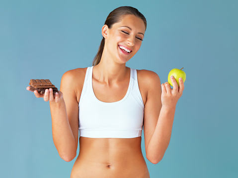 Studio shot of a fit young woman deciding whether to eat a chocolate or an apple against a blue background