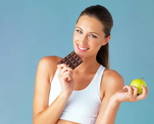 Studio shot of a fit young woman holding an apple and a chocolate against a blue background