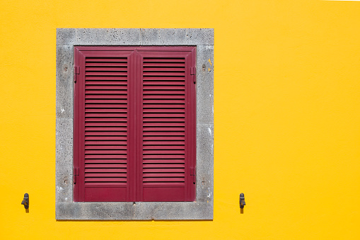 Bright yellow facade with a stone framed grey window, covered with red shutter.