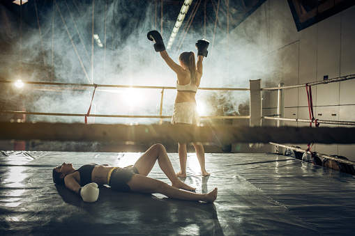 Back view of a female boxer celebrating victory after knocking out her opponent in a ring.
