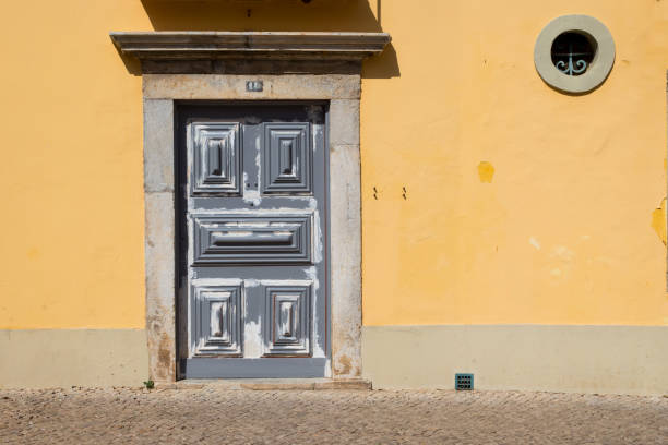 Closed grey door of an old yellow house. Round small window. Street full of sunshine. Faro, Portugal.