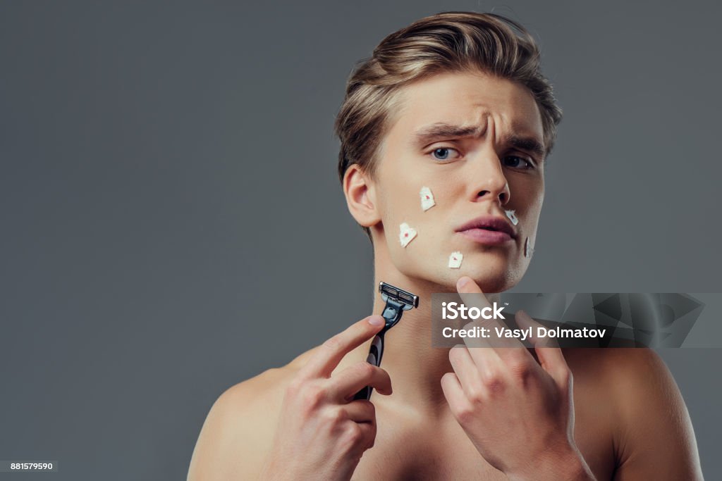 Handsome man on grey background. Handsome young man isolated. Portrait of shirtless muscular man is standing on grey background with razor in hand while shaving. Men care concept. Get hurt while shaving. Shaving Stock Photo