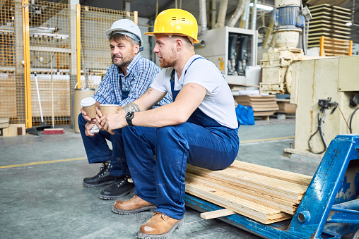 Full length portrait of two builders wearing hardhats taking break from work drinking coffee and resting sitting on pack of wood indoors