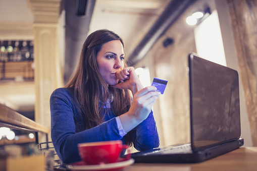 Young woman drink a cappuccino at the cafe, using laptop for online card payment. She is worried