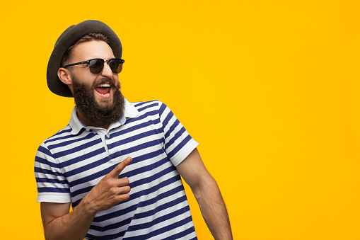 Young bearded hipster in cap and sunglasses pointing happily away on orange background.