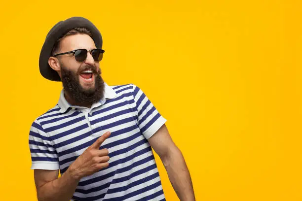Young bearded hipster in cap and sunglasses pointing happily away on orange background.