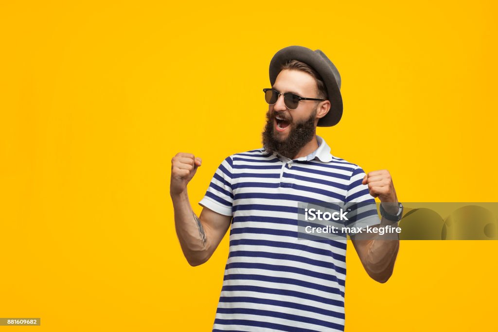 Excited man looking victorious Young hipster man in sunglasses and hat holding fists up celebrating victory on orange background. Colored Background Stock Photo