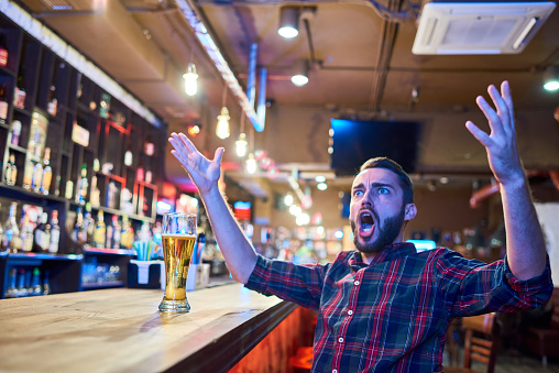 Portrait of bearded sports fan cheering emotionally in pub watching  match on TV sitting at bar counter with hands raised in astonishment, copy space