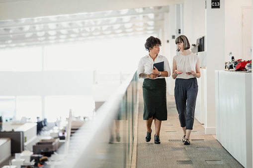 Two female professionals having a relaxed meeting while strolling along a raised walkway in an office auditorium.